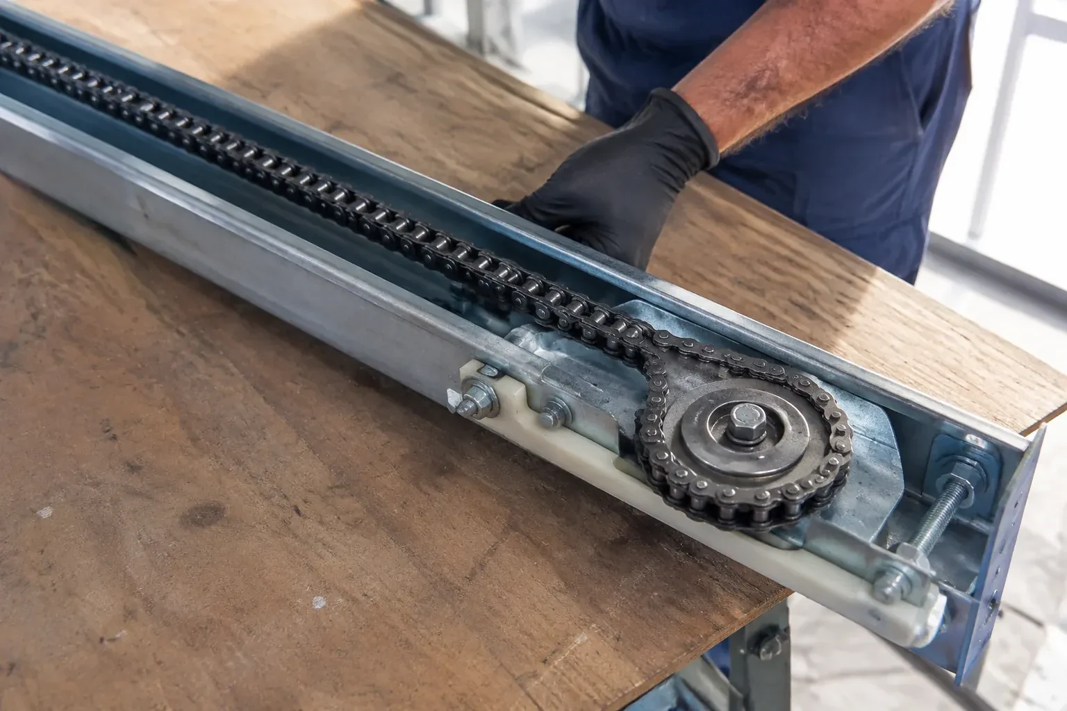 Gloved hand resting on a steel opener rail laid on a wooden bench, showing a drive chain looped around a sprocket wheel. — Chain-Drive Opener Repair, Markham
